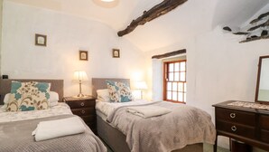 A room with two beds, each with a nightstand, a lamp, and a window. There is a dresser with a mirror and a small table with a lamp. - Castle Lodge (Keswick)