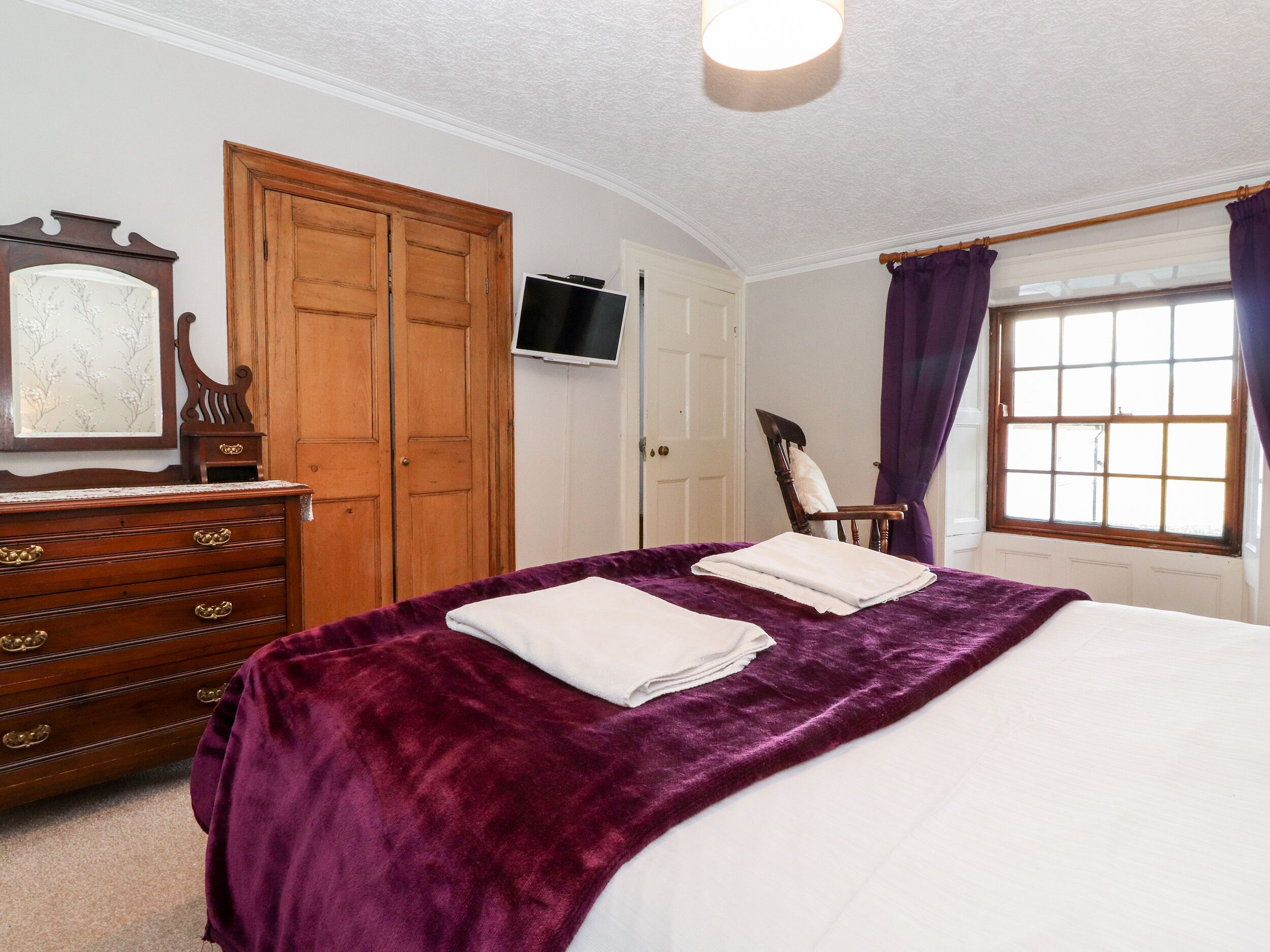 A bedroom with a bed, a wooden dresser, a television, and a window with curtains.