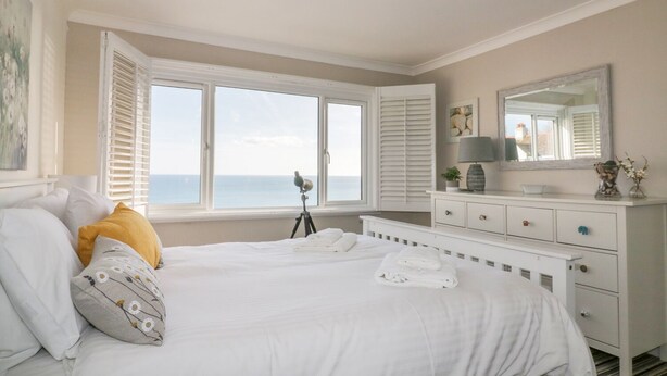A bedroom with a bed, a dresser, a mirror, and a window with shutters. - Bayview (St Austell)