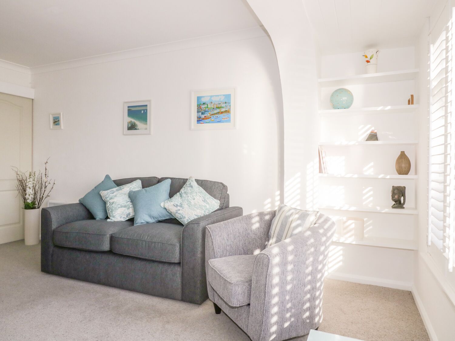 A living room with a grey sofa, a patterned armchair, and a white shelving unit with decorative items.