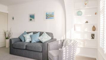 A living room with a grey sofa, a patterned armchair, and a white shelving unit with decorative items.