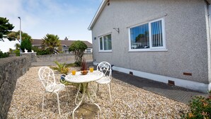 Ein Haus mit einer Terrasse, einem Tisch und Stühlen, einem kleinen Garten und blauem Himmel.