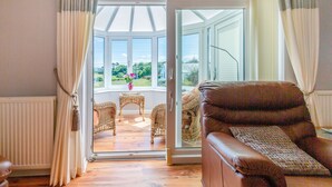 Ein Wohnzimmer mit einem braunen Ledersofa, Korbstühlen und einer Glastür, die auf einen Balkon mit Blick auf Grünflächen führt.