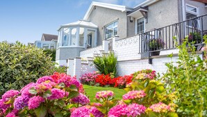 Ein modernes Haus mit weißer Fassade, einem Balkon mit schwarzen Geländern und einem Garten mit verschiedenen Blumen.