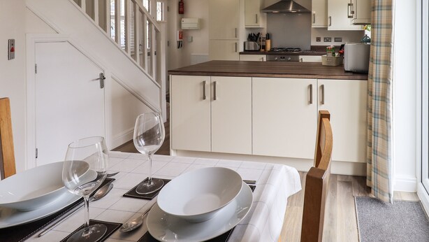 A modern kitchen with white cabinets, a wooden countertop, and a dining area set with plates, glasses, and cutlery. - Shore View (Grange-over-Sands)