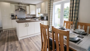 A modern kitchen with white cabinets, a wooden dining table set for two, and a view of the outdoors through large windows. - Shore View (Grange-over-Sands)