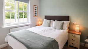 A neatly made bed with a white and green blanket, two bedside tables with lamps, a window with a view of trees, and a wall-mounted picture frame. - Shore View (Grange-over-Sands)
