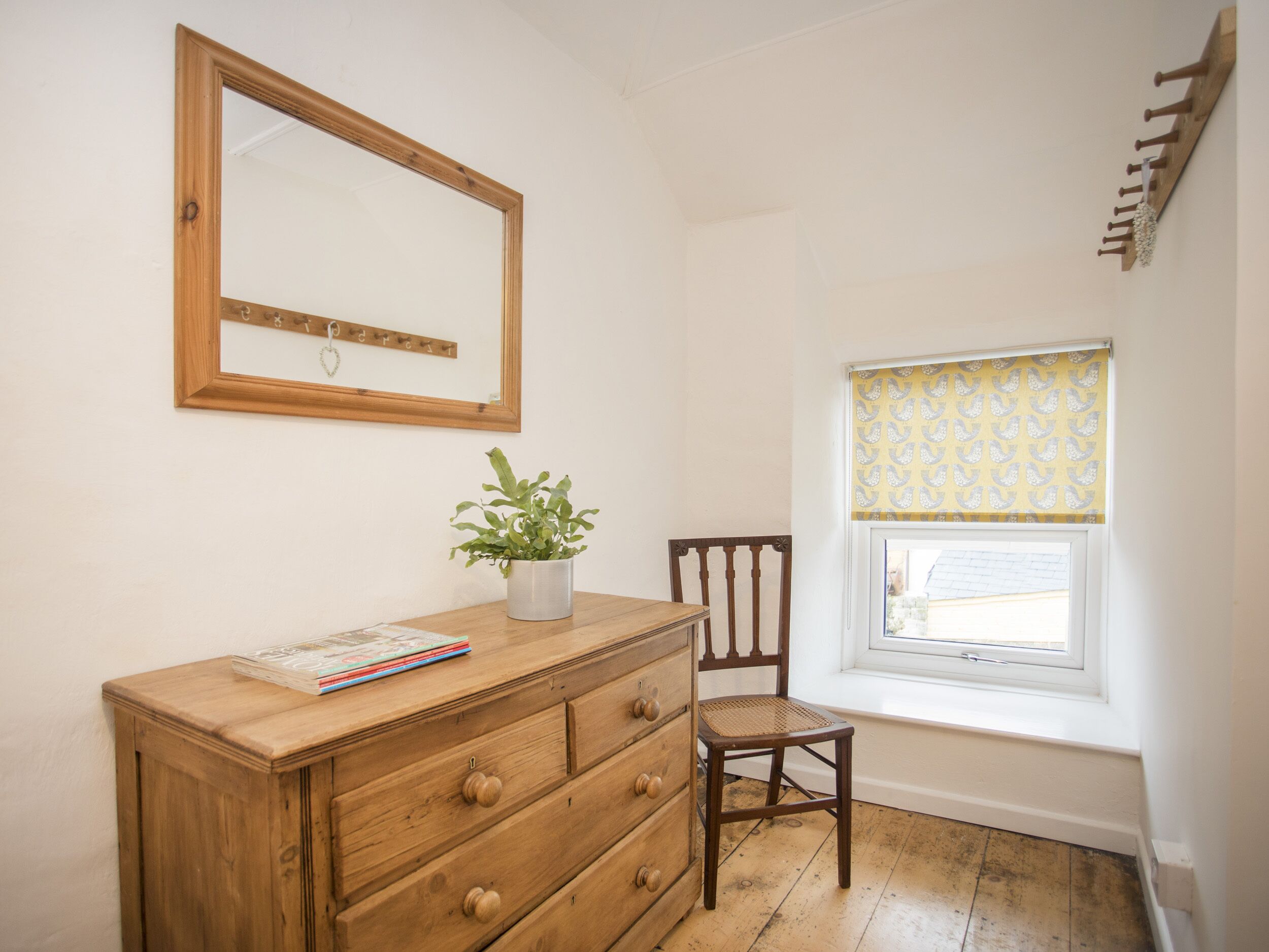 Een kleine kamer met een houten dressoir, een stoel, een potplant, een spiegel en een raam met decoratieve gordijnen.