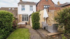 Une maison à deux étages, avec une façade en briques, une façade blanche et un petit jardin agrémenté d’une terrasse pavée et de mobilier d’extérieur.