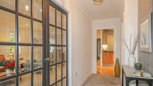 A hallway with a glass door leading to a living area, a wooden table with a vase, and a kitchen in the background.