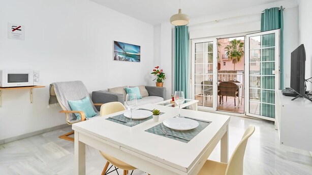 Un salon moderne comprenant un coin repas, un canapé, un téléviseur et un balcon offrant une vue sur les immebles et les palmiers.