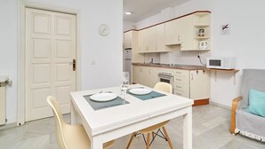 Une cuisine compacte avec des meubles blancs, un four à micro-ondes et une cuisinière. On y trouve une table à manger avec des chaises et une horloge au mur.