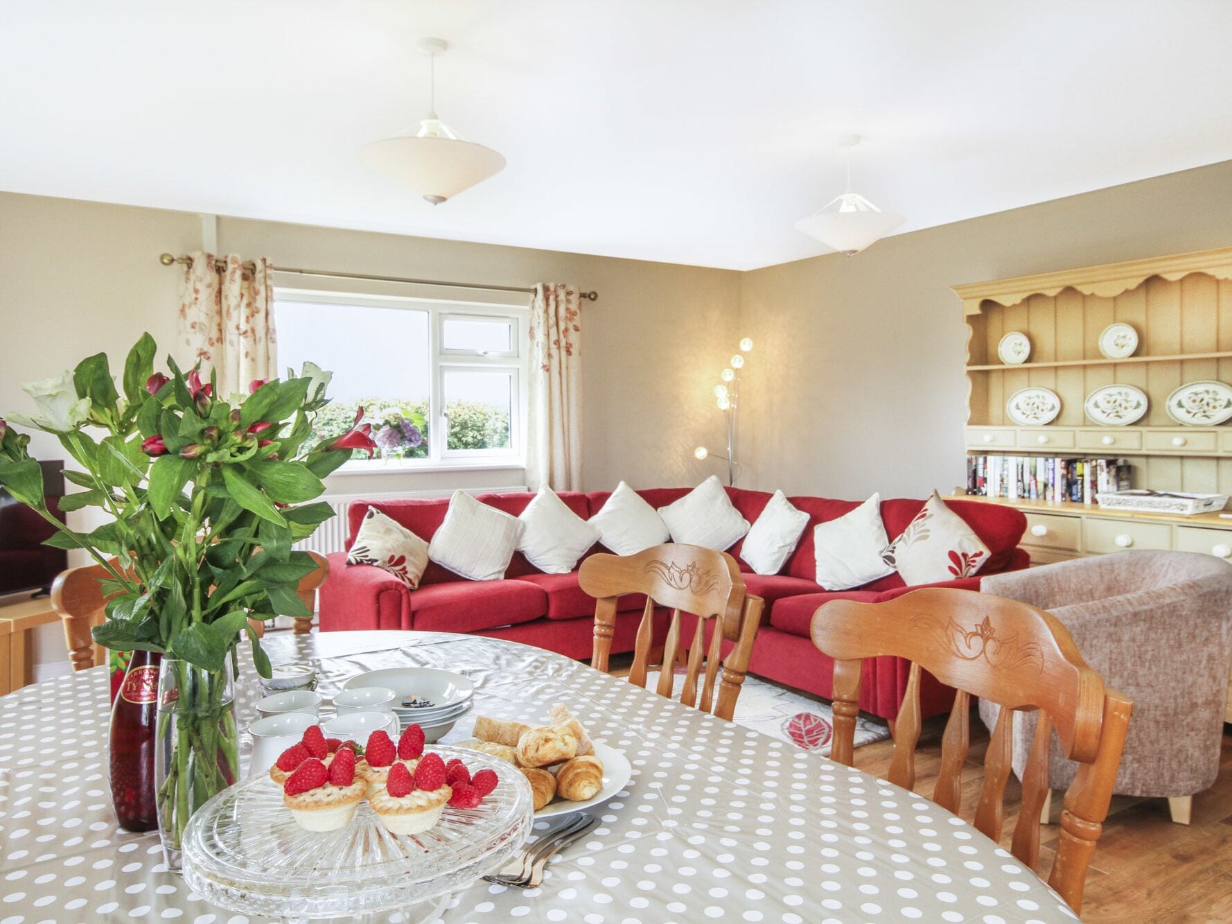 A cozy living room with a red sofa, wooden chairs, a dining table with a white polka dot tablecloth, and a vase of flowers.