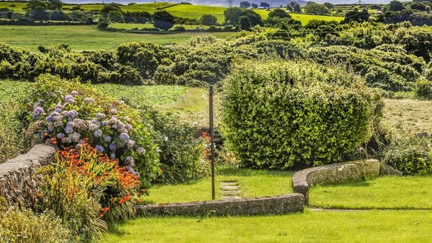 Paisagem rural com colinas ondulantes, um muro de pedra e diversa vegetação.