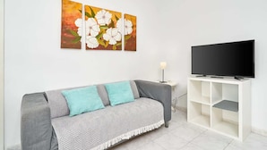 Ein Wohnzimmer mit grauem Sofa, einem Fernseher auf einem weißen Schrank und drei floralen Bildern an der Wand.