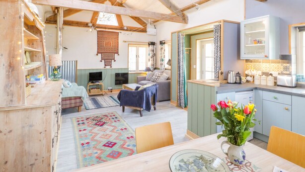 Un interior acogedor y rústico con vigas de madera a la vista, una zona de comedor con mesa y sillas, una cocina con armarios y una sala de estar con sofá y televisor.