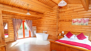 Ein Schlafzimmer in einer Blockhütte mit einem hölzernen Bett, einem roten Teppich mit Blumenmuster, einer weißen Badewanne und einem Blumenbild an der Wand.