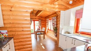 Ein Holzblockhaus-Interieur mit Holzboden, einem Essbereich mit Tisch und Stühlen sowie einer Küche mit weißen Schränken und einer roten Fensterkuche.