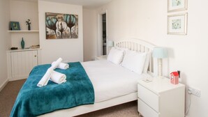 A neatly made bed with a teal blanket and rolled towels, a white nightstand with a lamp, and a painting on the wall. - Firkin Cottage (Weymouth)