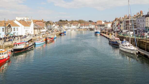 A harbor with boats docked along the waterfront, surrounded by buildings with various architectural styles. - Firkin Cottage (Weymouth)