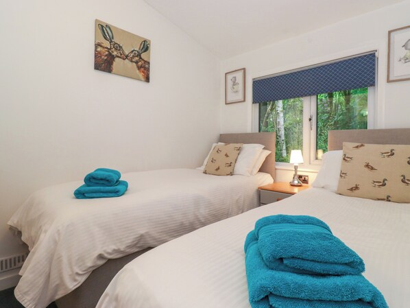 A hotel room with two beds, each with a white bedspread and matching pillows. There is a window with a blue blind, a bedside table with a lamp, and a framed picture on the wall. - 40 Skiptory Howe (Windermere)