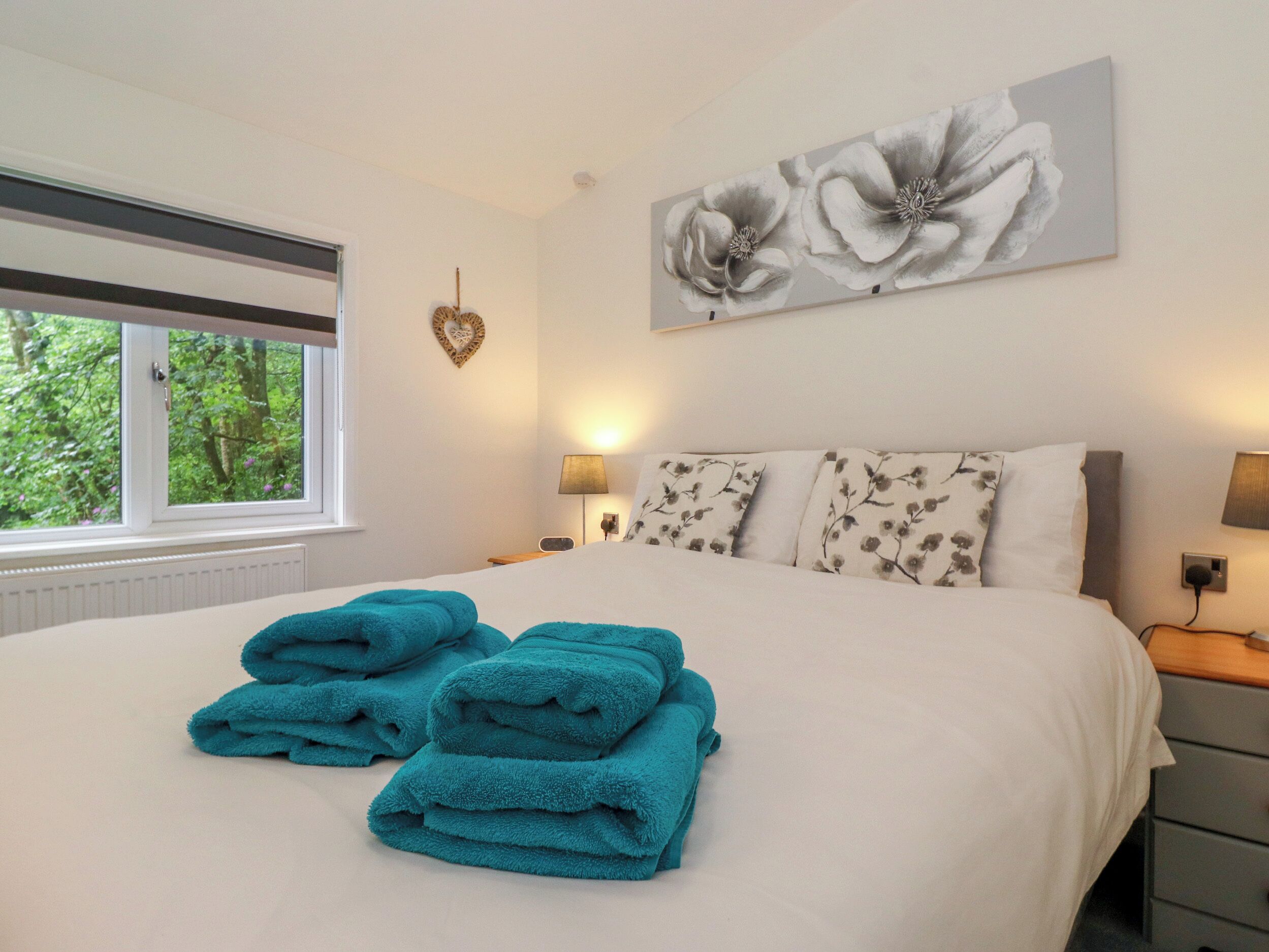 Une chambre avec un grand lit, une fenêtre donnant sur un paysage verdoyant, une décoration en forme de cœur et un tableau floral accroché au mur.