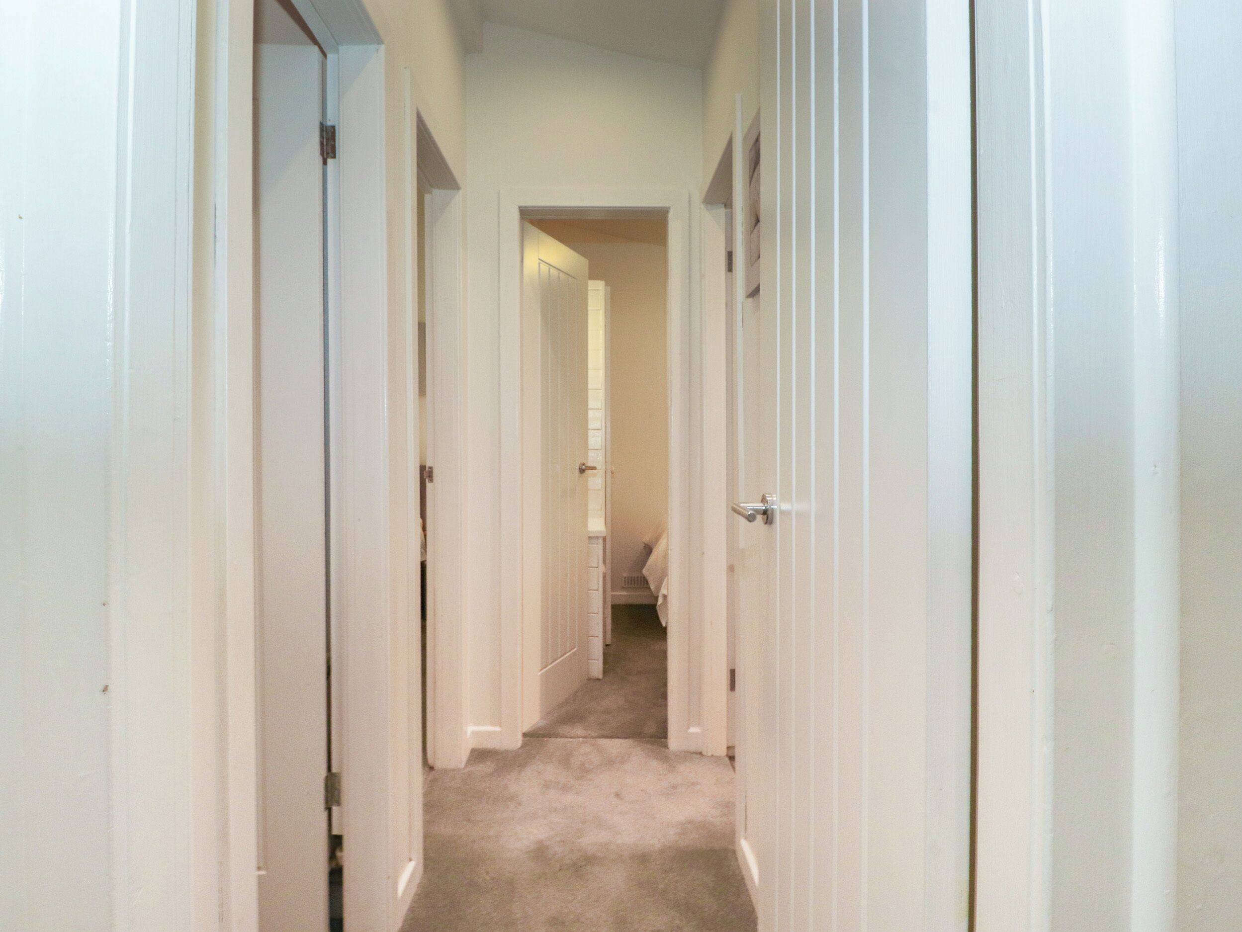 A narrow hallway with white doors on both sides, leading to a room with a visible bed and a small window.