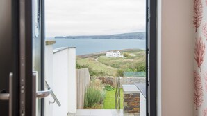 Vue depuis une pièce dotée d’une porte vitrée, donnant sur un paysage côtier avec un bâtiment blanc et une étendue d’eau.