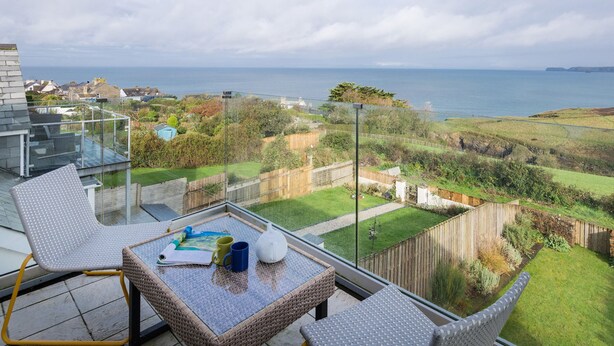 Un balcon doté d’une paroi vitrée donnant sur un paysage côtier, avec une table en osier sur laquelle sont posés un livre, une tasse et une théière.
