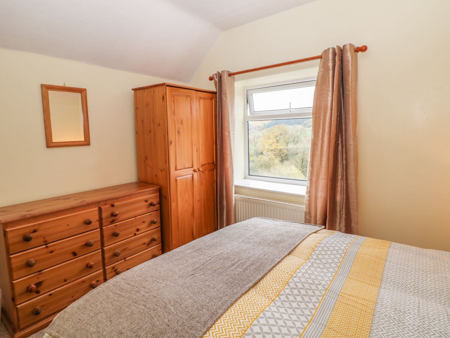 A bedroom with a wooden dresser, a bed with patterned bedding, a window with curtains, and a radiator.