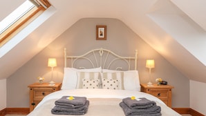 Quarto com teto inclinado, cama com roupa de cama branca, duas mesas de cabeceira com luminárias e um claraboia.