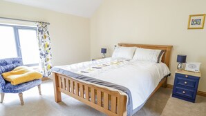 A bedroom with a wooden bed, a chair with a yellow blanket, a nightstand with a radio, and a window with curtains. - 7evern @ Porth Farm (Caersws)