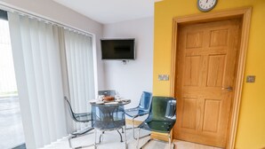 A room with a wooden door, a flat-screen TV mounted on the wall, a table with chairs, and a window with blinds. - 7evern @ Porth Farm (Caersws)