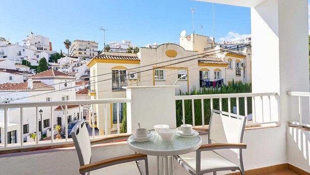 Un balcon avec une table et des chaises, donnant sur un quartier résidentiel avec des bâtiments blancs et un ciel dégagé.