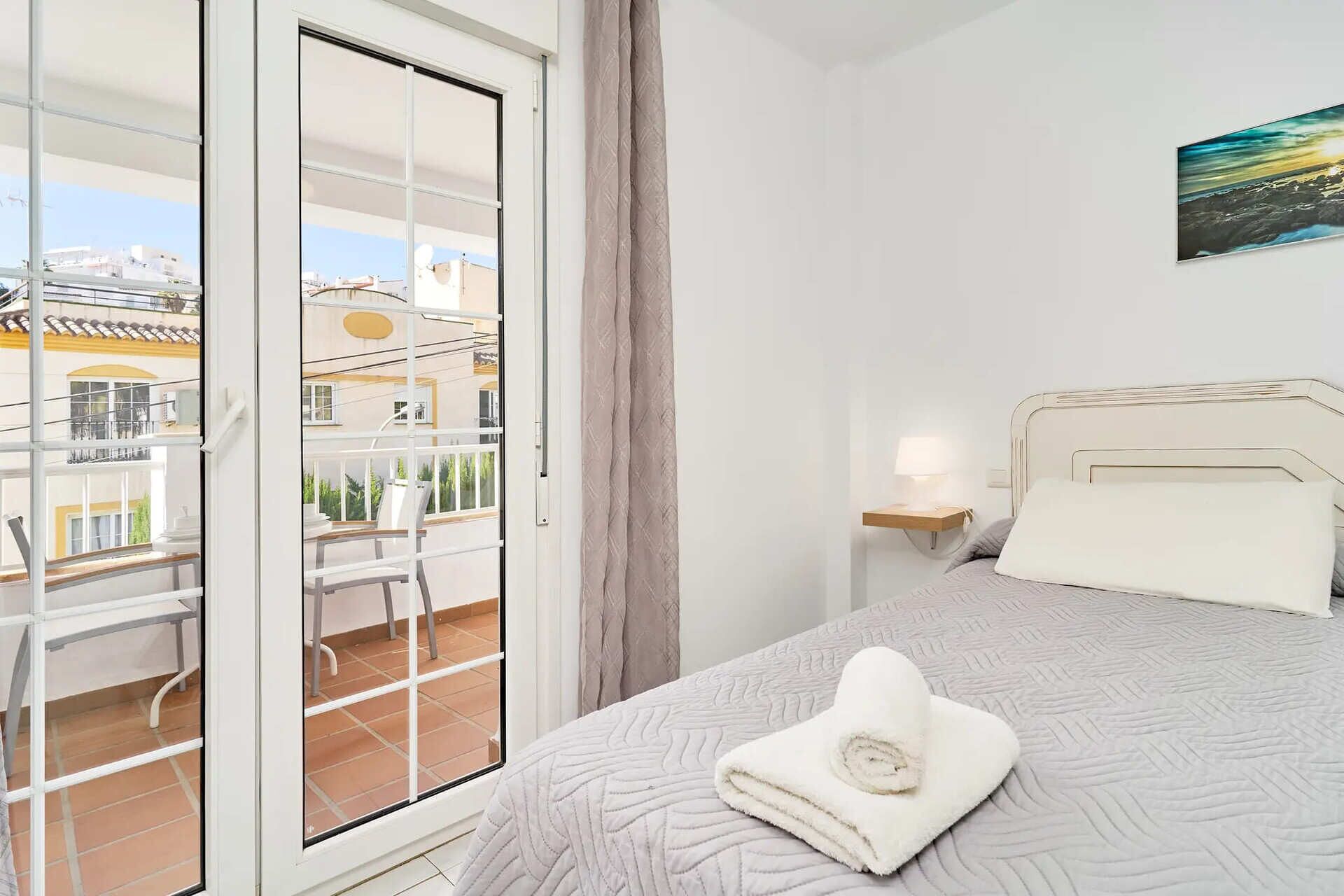 A bedroom with a bed, a nightstand, a window with a view of buildings, and a balcony with a chair.