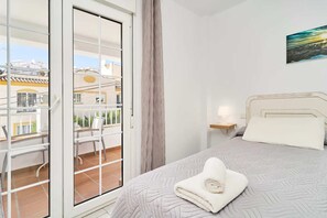 A bedroom with a bed, a nightstand, a window with a view of buildings, and a balcony with a chair. - Suite Nº 8 (1 double room) (Nerja)