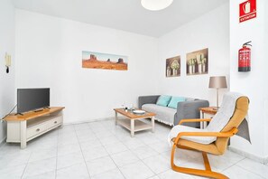 Una sala de estar moderna con un televisor de pantalla plana, un sofá, una mesa de centro y una silla. Hay pinturas en la pared y un extintor colgado en la pared.