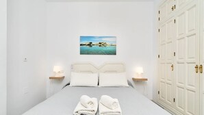 A neatly made bed with two pillows, a bedside table with a lamp, a painting of a landscape on the wall, and a large wardrobe with double doors. - BB Suite (3 double rooms) (Nerja)