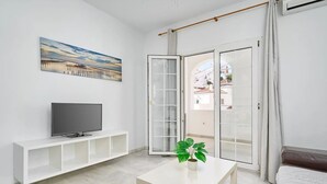 A modern living room with a flat-screen TV on a white console, a painting above the TV, a sliding glass door leading to a balcony, and a potted plant on a table. - BB Suite (3 double rooms) (Nerja)