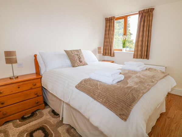 A neatly made bed with white linens, a brown throw blanket, and a decorative pillow. There is a wooden nightstand with a lamp and a drawer. A window with curtains is visible, and a radiator is on the wall. - Stonecroft Lodge (Belper)