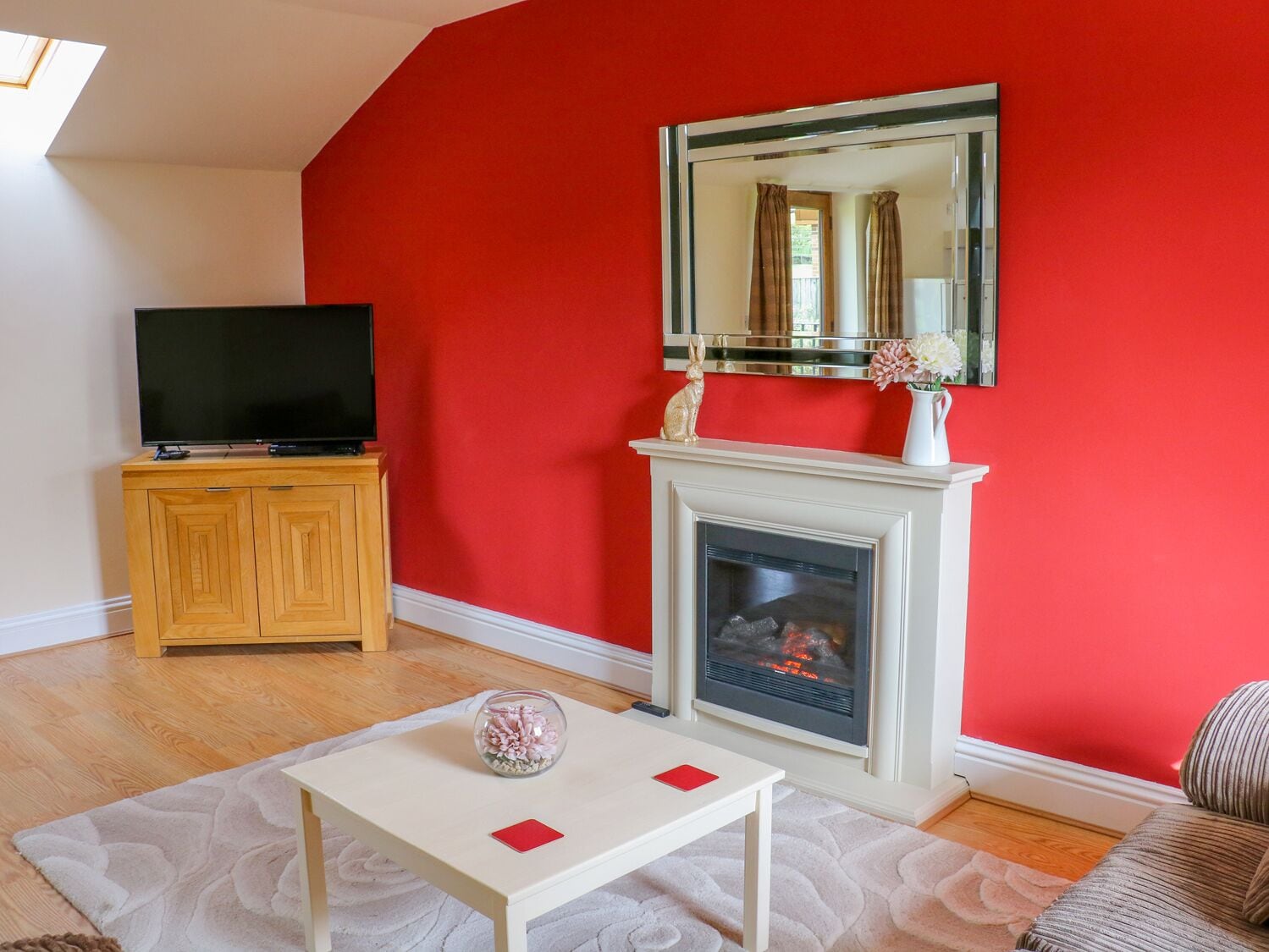 Un salón con una pared roja, una chimenea, un televisor sobre un mueble de madera y una mesita con un jarrón y un libro rojo.