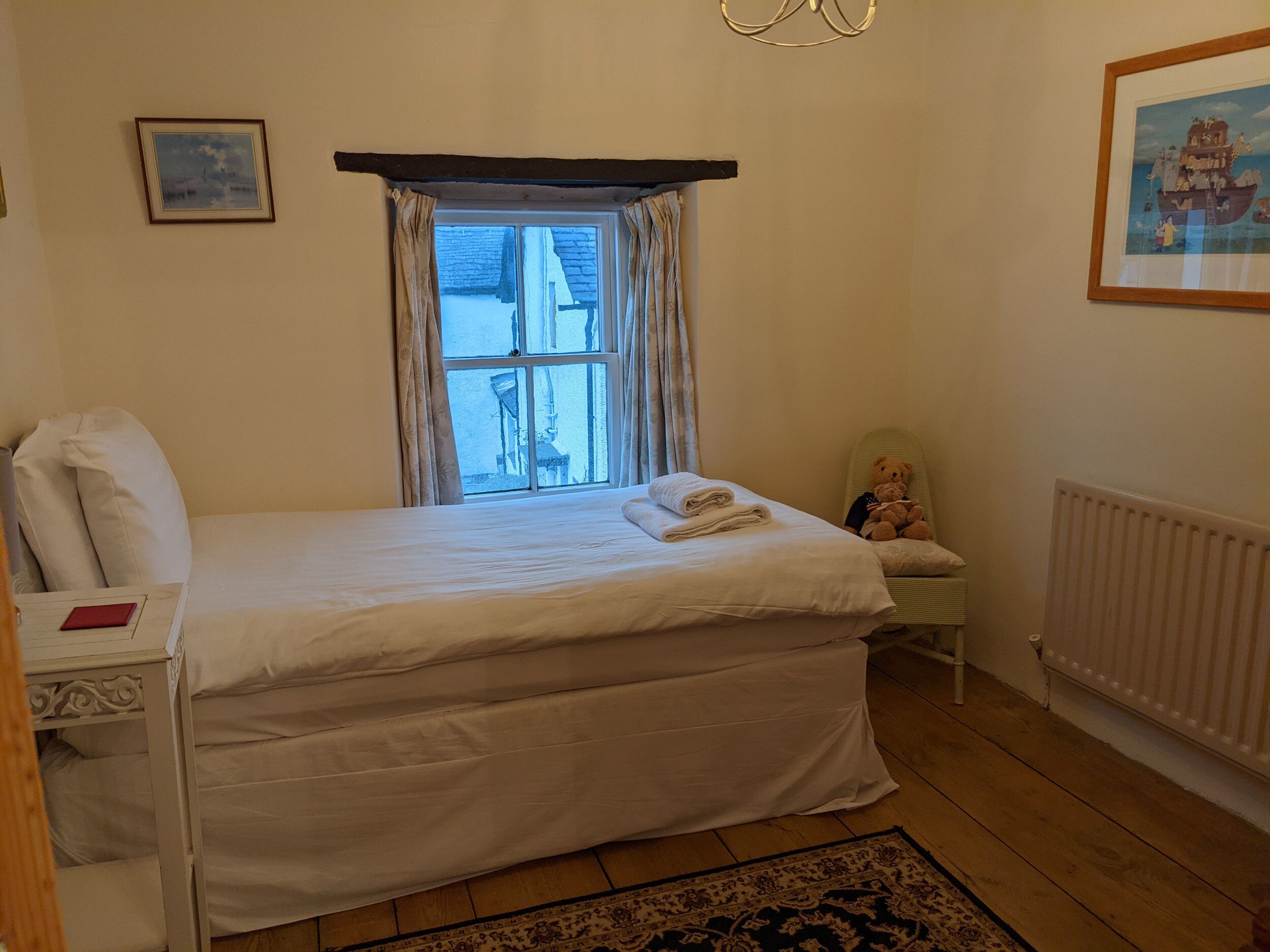 Une chambre à coucher avec un lit, une fenêtre avec des rideaux, un ours en peluche, un radiateur et un tableau encadré accroché au mur.