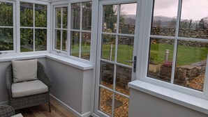 Un invernadero con ventanas de marco blanco, mobiliario de mimbre y vistas a un jardín con un muro de piedra.