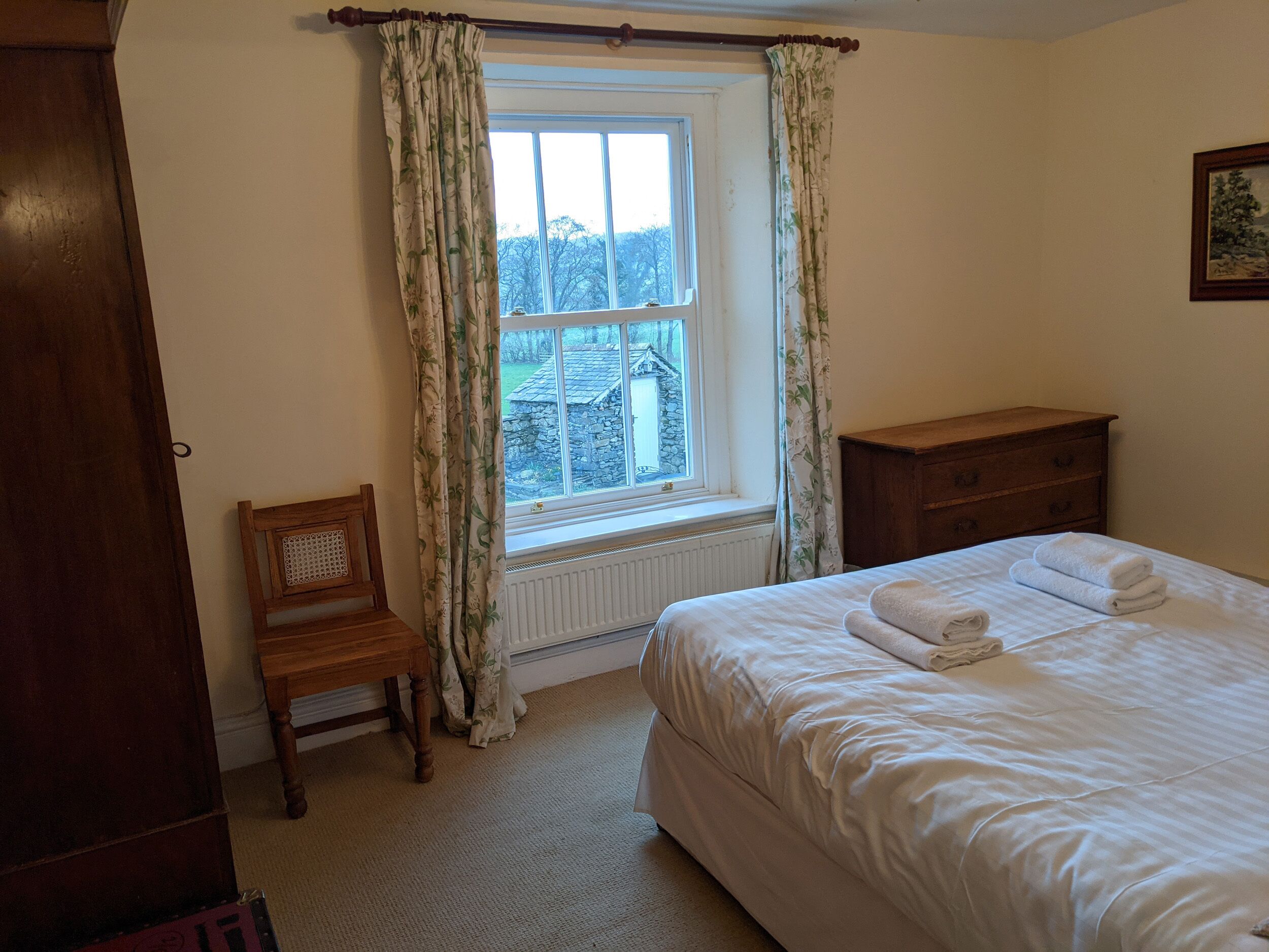 A bedroom with a bed, a wooden nightstand, a chair, and a window with curtains.