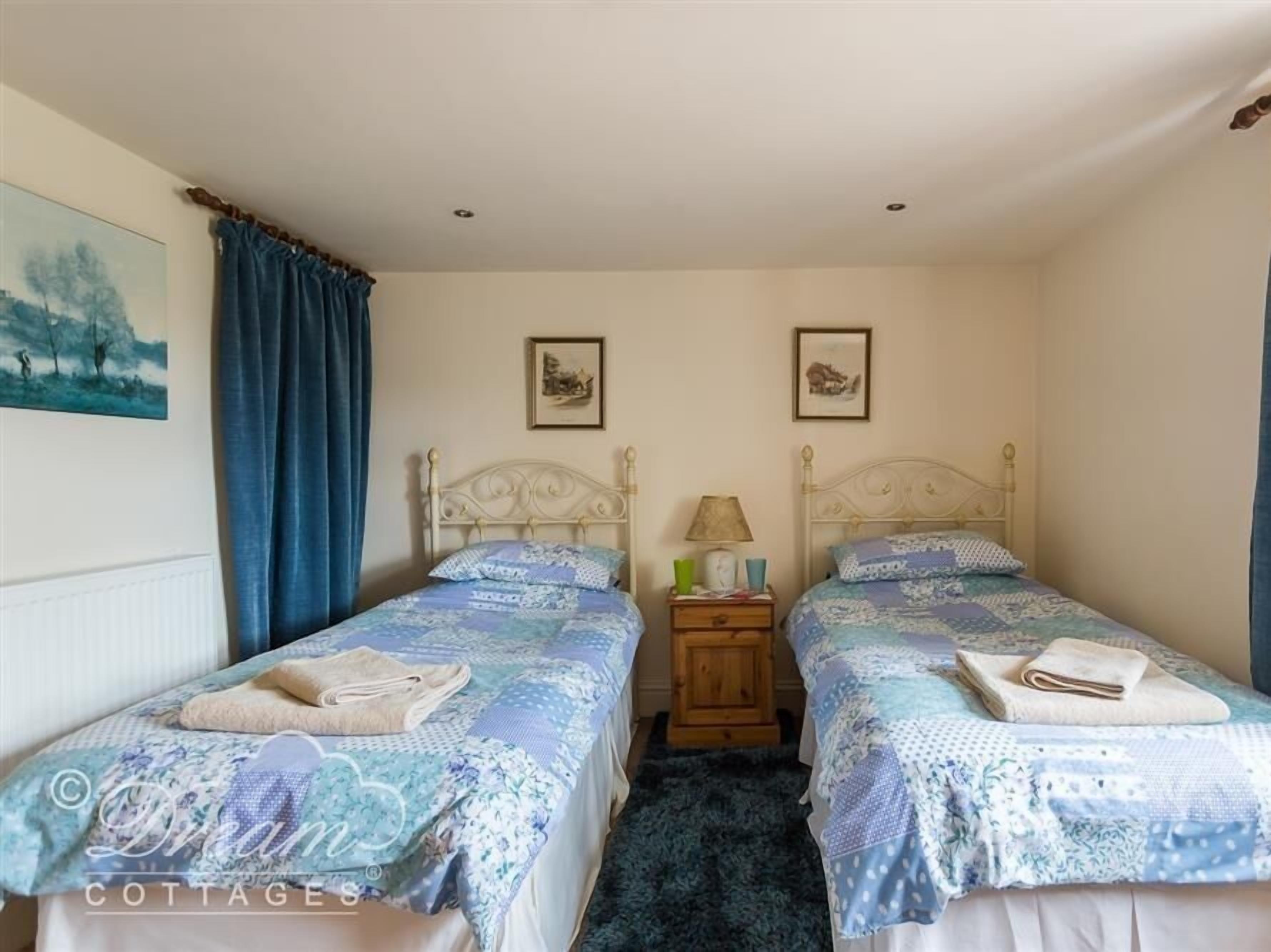 A bedroom with two single beds, a wooden nightstand, a lamp, and two framed pictures on the wall.