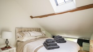 Ett sovrum med ett sluttande tak, en säng med en beige sänggavel, en sänglampa och en hög med hopvikta handdukar.