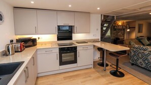 A modern kitchen with white cabinets, a built-in oven, a microwave, and a toaster. There is a bar stool and a patterned sofa in the adjacent living area. - Orchard Cottage (Whitby)