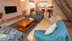 A cozy living room with a sofa, a coffee table, and a patterned armchair. A TV is mounted on the wall, and there's a small kitchen area with a sink and a microwave. - Orchard Cottage (Whitby)