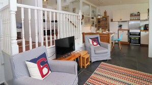 Un vaste espace de vie comprenant un téléviseur, deux fauteuils à rayures, une table en bois et, au fond, une cuisine.