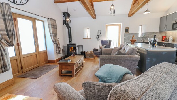 A spacious living room with a fireplace, wooden beams, a sofa, a coffee table, and a kitchen area. - Apple Tree Cottage (Appleby-in-Westmorland)